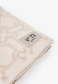 Textil beige con un patrón geométrico texturizado, que presenta una etiqueta plateada que dice "V°73 VENEZIA". La tela parece suave y esponjosa.