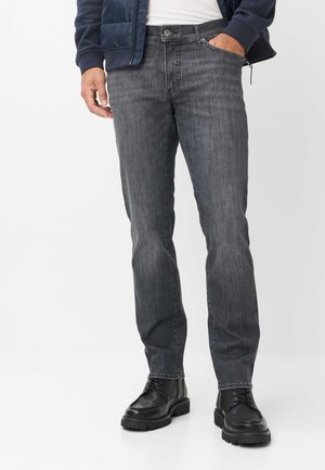Mand iført mørkegrå jeans, sorte snørebåndsstøvler, hvid skjorte og navyblå jakke med den ene hånd i lommen, der står på en hvid baggrund.
