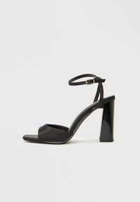 Μαύρο σανδάλι με ψηλοτάκουνο που διαθέτει υφασμένη λουρά με πόρπη, σχέδιο ανοιχτής μύτης και γυαλιστερό τετρά�γωνο τακούνι. Ομοιόμορφο λείο υλικό σε όλο το παπούτσι.