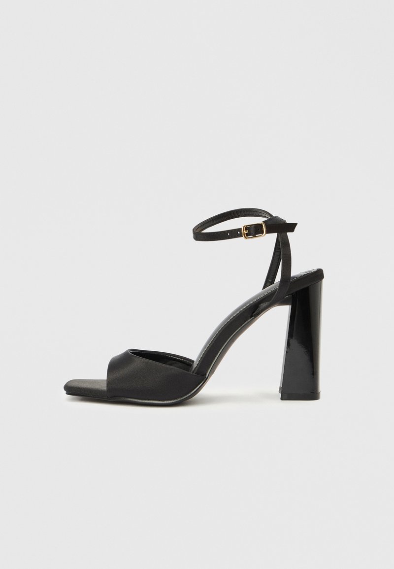 Μαύρο σανδάλι με ψηλοτάκουνο που διαθέτει υφασμένη λουρά με πόρπη, σχέδιο ανοιχτής μύτης και γυαλιστερό τετράγωνο τακούνι. Ομοιόμορφο λείο υλικό σε όλο το παπούτσι.