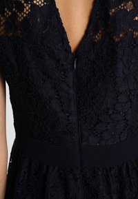 Svart spetsklänning med blommönster, djup V-ringning, figurnära midja och genomskinlig spets på övre bröstet och axlarna.