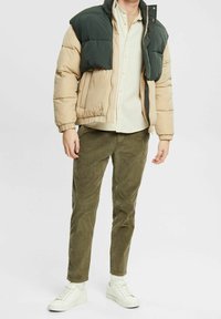 Giacca a vento con parte superiore verde e parte inferiore beige, abbinata a pantaloni in corduroy oliva e scarpe da ginnastica bianche. Tessuto liscio, chiusura con zip.