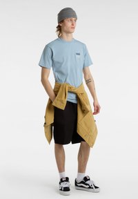 Světle modré tričko s logem Vans, černé cargo šortky, žlutá tkanina uvázaná v pase, šedý beanie a černé tenisky Vans. Hladké textury.