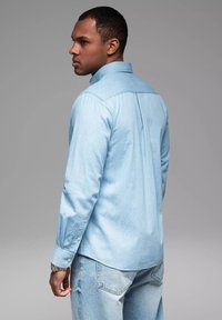 Lichtblauwe blouse met lange mouwen, een gestructureerd patroon, een button-down kraag en een contrasterende achterbies. Gecombineerd met versleten denim jeans.