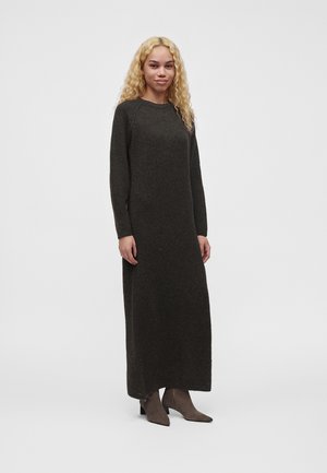 Femme aux cheveux blonds bouclés portant une longue robe en maille gris foncé et des bottines marron, debout devant un fond clair uni.