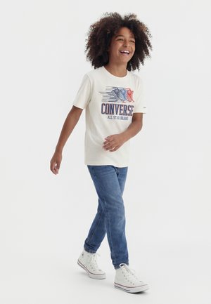 Enfant souriant aux cheveux bouclés marchant, portant un t-shirt graphique blanc Converse, un jean bleu et des baskets montantes blanches sur un fond uni.