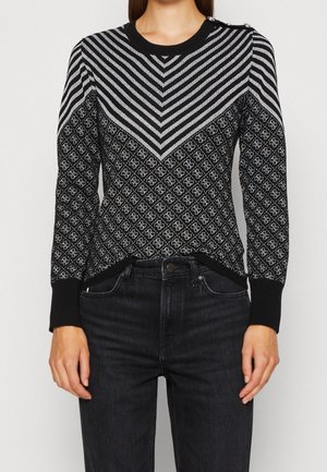 Frau trägt einen schwarzen und grauen Langarm-Pullover mit Chevron-Streifen und Rautenmuster, kombiniert mit hoch taillierten schwarzen Jeans.