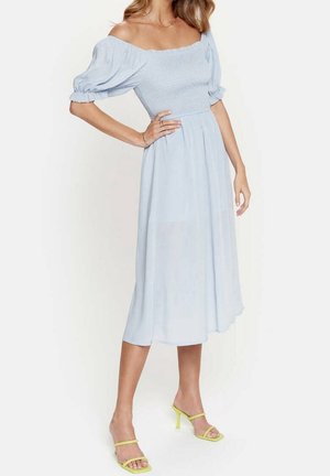 Frau trägt ein hellblaues Off-Shoulder-Midikleid mit Puffärmeln und gelbe Sandalen mit Absatz, posiert mit einer Hand auf der Hüfte.