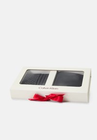Conjunto de presente Calvin Klein numa caixa branca com laço vermelho; inclui uma carteira preta texturizada e um porta-cartões preto liso, visíveis através da janela.