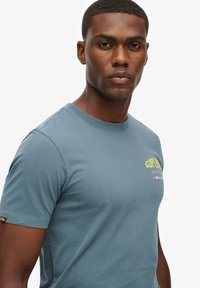 Camiseta de algodón azul con mangas cortas, escote redondo y estampado de logotipo en amarillo. Textura suave y diseño ajustado.