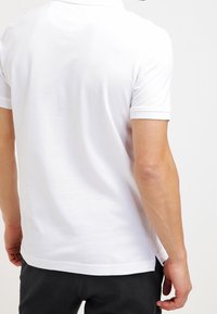 Camisa polo blanca con un cuello clásico y mangas cortas, mostrada desde la parte trasera, con una textura suave y aberturas en los laterales del dobladillo.