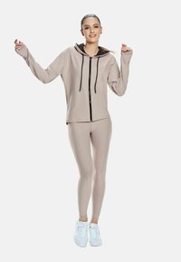 Young woman standing, wearing beige zip-up hoodie, matching leggings, and white sneakers, with arms raised and smiling.