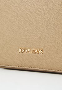Bež torba iz teksturirane usnje s gladko obdelavo, ki ima kovinski zlat "JOOP! JEANS" logo in diskretne šivne detajle ob robu.