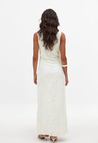 Pitkä, valkoista virtaava mekko, jossa on kukkatekstuuri, olkaimeton muotoilu ja hieman istuva vyötärö. Yhdistetty valkoisiin hihnakenkiin.