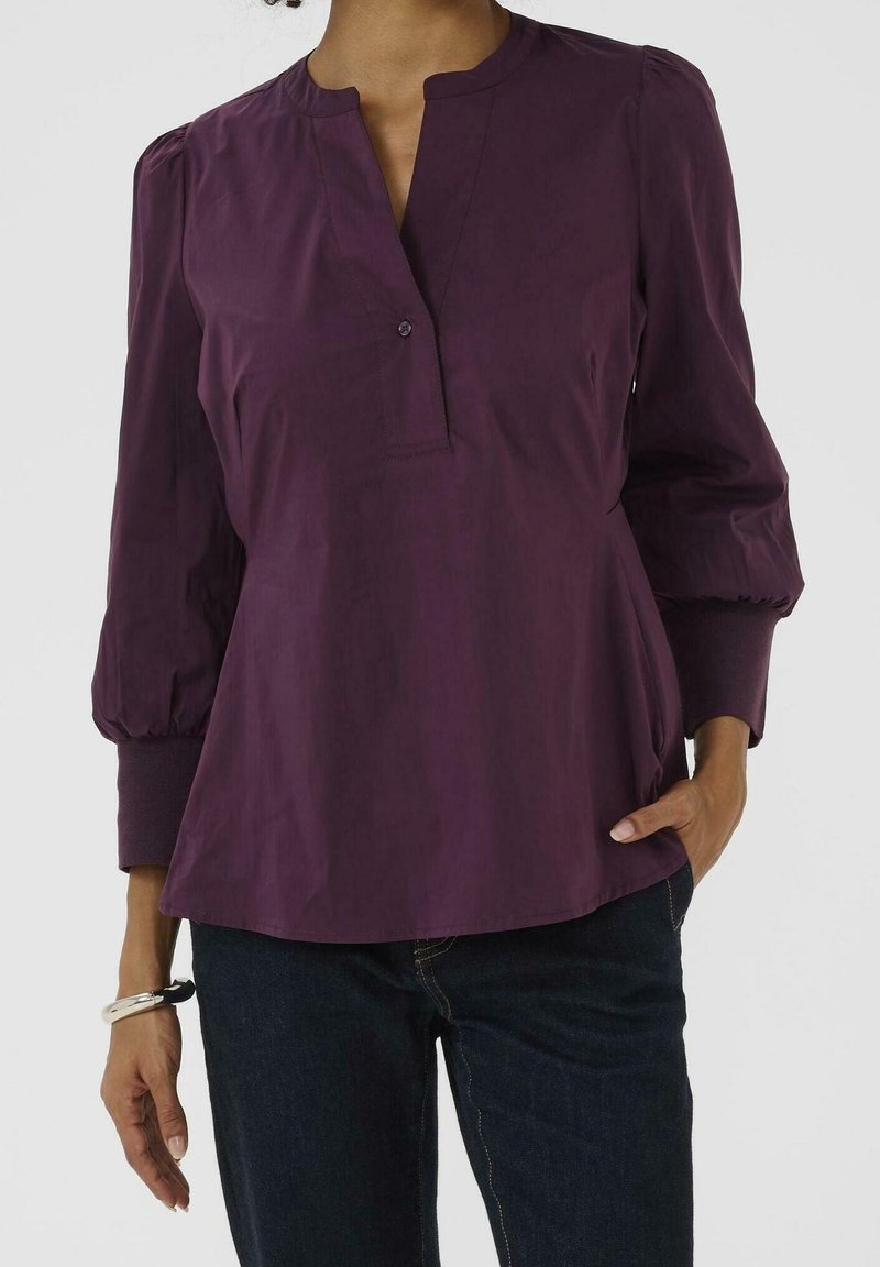 Femme portant un chemisier violet à manches longues avec des manches à revers et une encolure à un seul bouton, associée à un jean bleu foncé et un bracelet argenté.