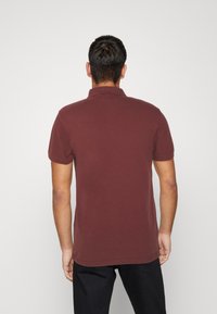 Homme de dos portant un polo à manches courtes maron et un pantalon noir sur un fond blanc uni.