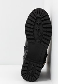 Černá podrážka boty s robustním vzorem tread, obsahující hluboké drážky a vyvýšené akcenty. Hladký horní materiál viditelný na okraji.