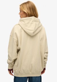 Vrouw met blond haar die een oversized beige hoodie en een blauwe spijkerbroek draagt, gezien van achteren tegen een witte achtergrond.