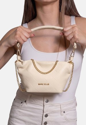 Woman wearing white tank top and pants holds beige Marc Ellis handbag with gold chain strap in front of her torso.