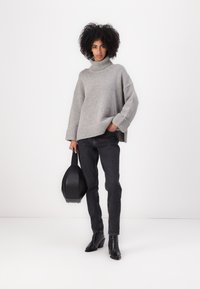 Jersey gris, de gran tamaño, con cuello de tortuga y detalles de canalé, combinado con jeans oscuros y botines negros, sosteniendo un bolso negro.