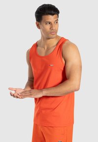 Pomarańczowy bawełniany top na ramiączkach z szerokimi ramiączkami, okrągłym dekoltem i małym logo na piersi. Modelka pozująca z wyciągniętymi rękami.