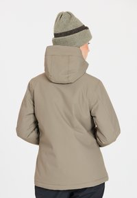 Beige isolerad jacka med huva, visad från baksidan. Har en slät yta och minimalistisk design. Buren med en grönrandig stickad mössa.