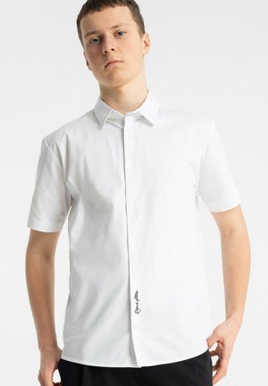 Młody mężczyzna w białej, krótkorękawowej koszuli zapinanej na guziki z czarnym napisem przy dolnym guziku, stojący na tle jednolitego jasnego tła.