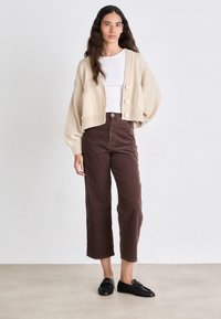 Beige cropped cardigan met grote knopen, gecombineerd met bruine wijde corduroy broeken. Model draagt een witte shirt en zwarte instappers.
