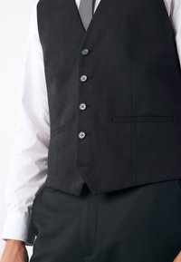 Gilet en tissu noir avec une finition texturée, comprenant six boutons, deux poches avant, et une coupe cintrée, porté sur une chemise blanche.