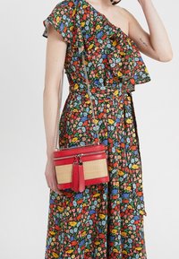 Czerwona i beżowa torebka na ramię z fakturowanym, plecionym przodem, srebrnym łańcuszkowym paskiem i ozdobami w formie frędzli, trzymana przez osobę w kwiecistej sukience.