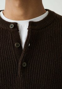 Brown ribbed knit pullover with a round neck and three-button placket, layered over a white crew neck shirt.