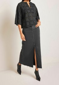 Femme portant un chemisier en dentelle œillet noir à manches trois-quarts, une jupe en denim foncé taille haute avec une fente frontale, et des bottes pointues noires.