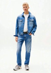 En blond mann iført blå denimjakke og jeans, hvit t-skjorte, svart belte og hvite sneakers, står med den ene hånden i lommen.