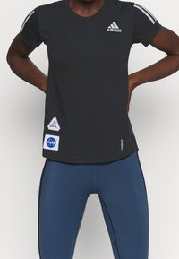 Černé tričko Adidas s bílým logem a nášivkami NASA. Má krátké rukávy, kulatý výstřih a volný střih, doplněné modrými legínami.