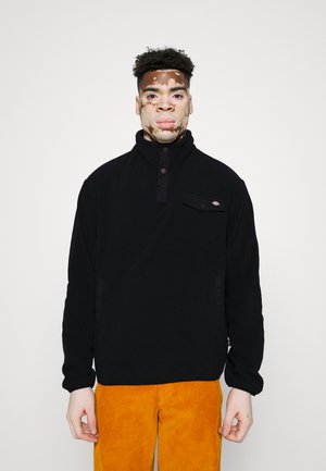 Pullover en polaire noir avec col montant, boutons-pression et poche poitrine. Le porteur l'associe à un pantalon en velours côtelé orange vif.