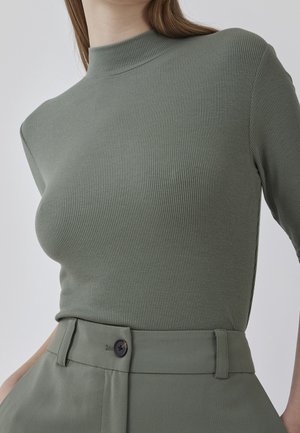 Vrouw draagt een olijfgroene geribde top met lange mouwen, ingestopt in een broek met hoge taille met een enkele zwarte knoop.