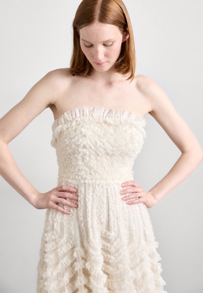Robe sans bretelles en crème avec une texture froncée, présentant un corsage ajusté et une jupe évasée avec des détails superposés. Tissu léger et lisse.