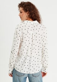Chemise blanche à manches longues avec col, arborant un motif de cœurs noirs. Tissu lisse, coupe légèrement loose et ourlet arrondi.