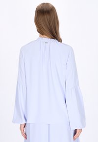 Lyseblå bluse med klokkeærmer, samlet halsudskæring og flydende design. Blødt stof med minimal hardware-detalje på bagsiden.