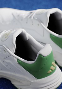Coppia di sneaker bianche con dettagli verdi sul tallone e lacci, poste su una superficie blu, mostrando i dettagli posteriori e laterali.