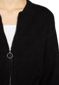 Musta neulottu neuletakki pyöreällä pääntiellä, täydellä vetoketjulla edessä ja hopeisilla yksityiskohdilla. Pehmeä pinta ja resorineuloskuvio.