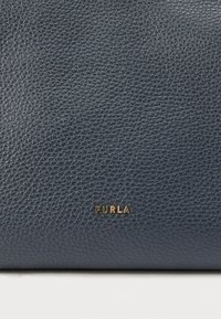 Tengerészkék bőr kézitáska, texturált felülettel, arany "FURLA" logóval. A dizájn letisztult, a minőséget és a kézművességet hangsúlyozza.