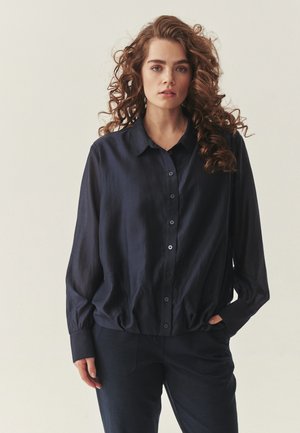 Młoda kobieta o kręconych włosach, ubrana w luźną, ciemnoniebieską koszulę na guziki i dopasowane spodnie, stoi na jasnym tle.