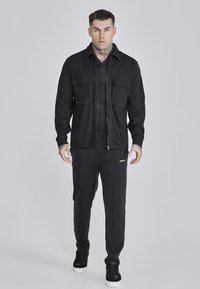Chaqueta negra con dos bolsillos en el pecho, cierre de cremallera y un acabado texturizado, combinada con pantalones ajustados a juego y zapatillas negras.