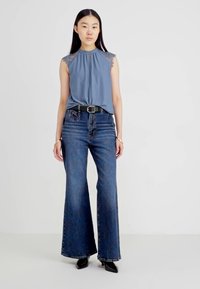 Top senza maniche blu chiaro con dettagli in pizzo, abbinato a jeans blu scuro a gamba larga e una cintura nera. Il modello si trova davanti a uno sfondo neutro.