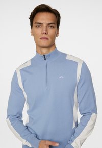 Ljusblå tröja med halvdax och hög krage. Har vita detaljer längs axlarna och ärmarna, tillverkad av ett mjukt stickat material.