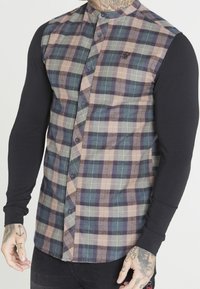 Camisa de manga larga con un frente de cuadros abotonado en verde, azul y beige, con mangas negras y un logo bordado en el pecho izquierdo.