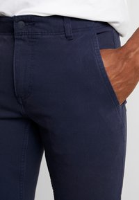 Námořnické modré bavlněné kalhoty s knoflíkovým zapínáním, bočními kapsami a hladkým povrchem, které zdůrazňují trvanlivou kvalitu látky.