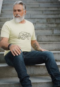 Light yellow t-shirt with a black geometric bear design, paired with dark denim jeans. Model seated on stone steps.