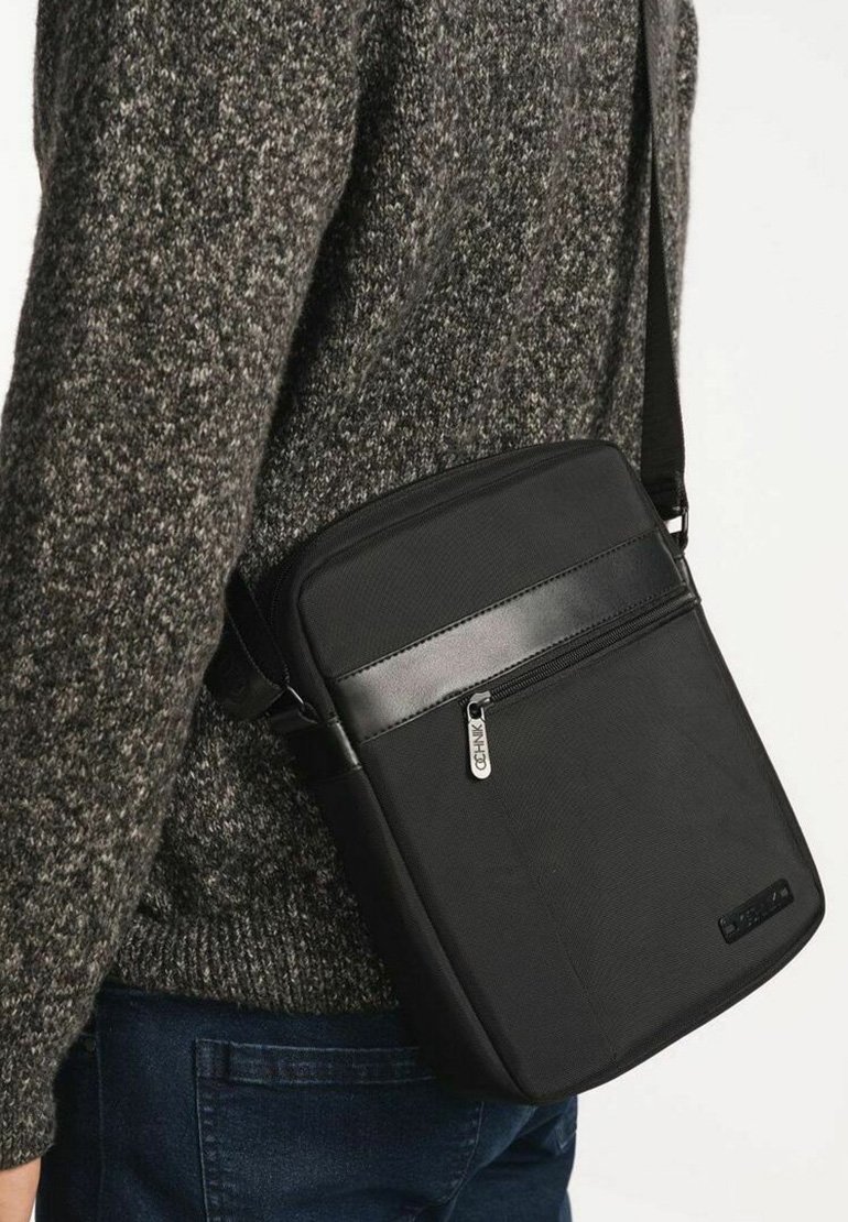 Czarna torba crossbody o gładkiej powierzchni, z przednią kieszenią na zamek i regulowanym paskiem, wykonana z trwałej tkaniny.